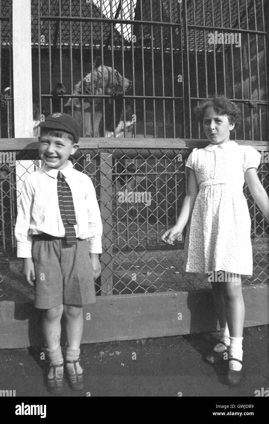1930s, historical, brother and sister in school uniform at a zoo, posing for a photograph in front of the bears in their cage. Stock Photo
