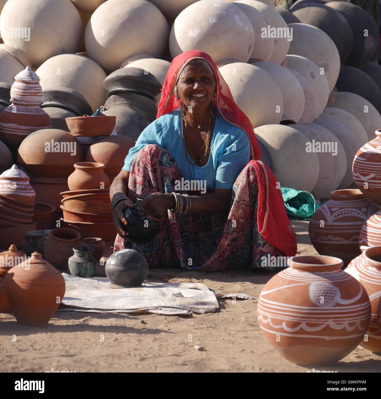 A smiling Indian woman in typical clothes and jewellery of Rajasthan, NW India sits on the ground selling round clay water pots Stock Photo