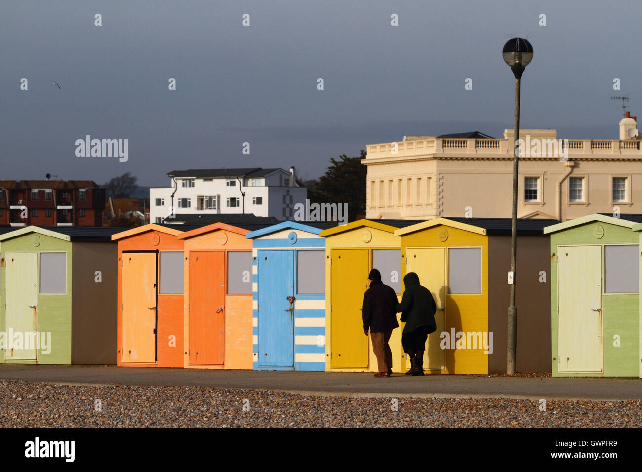 Elderly couple walking past line of wooden beach huts on sea front on a winter afternoon, Seaford, East Sussex, England Stock Photo
