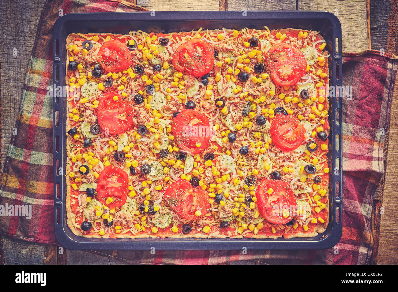 Vintage toned homemade tuna pizza with tomatoes, olives, onion and corn ready to bake on wooden background, top view. Stock Photo