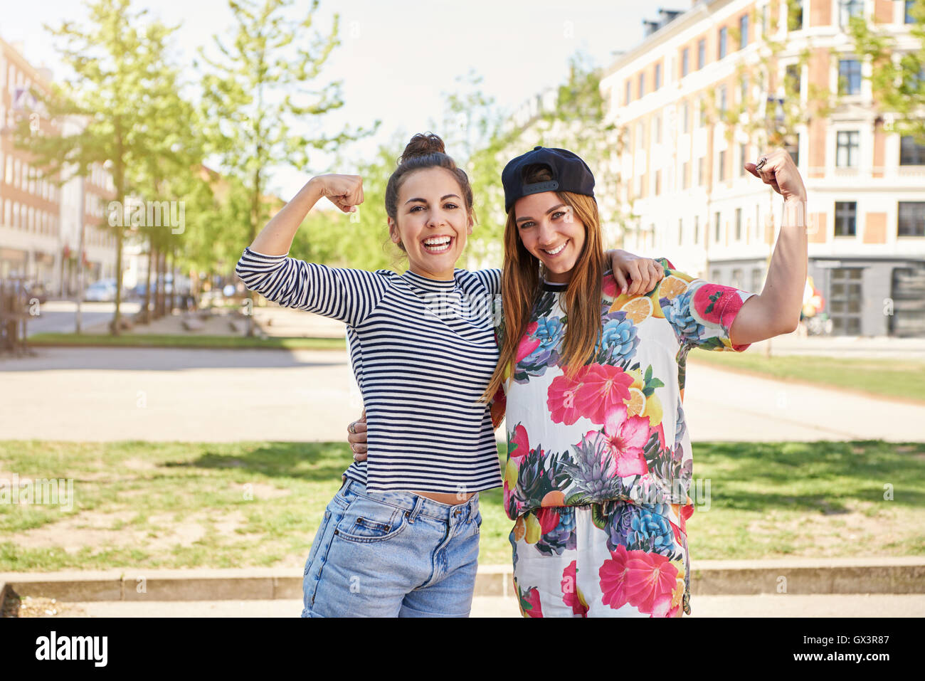 Cute vivacious young women friends standing arm in arm on a hot summer day in an urban street waving at the camera Stock Photo