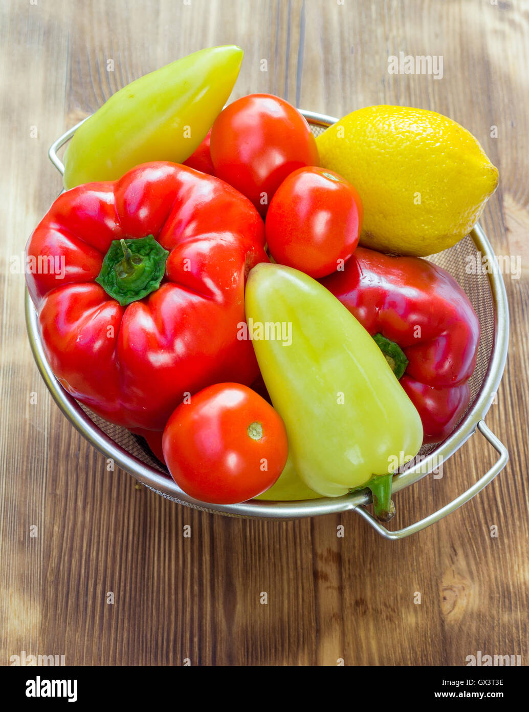 Vegetable still life of red and green peppers, tomatoes and lemon in sieve bowl on wooden board background Stock Photo