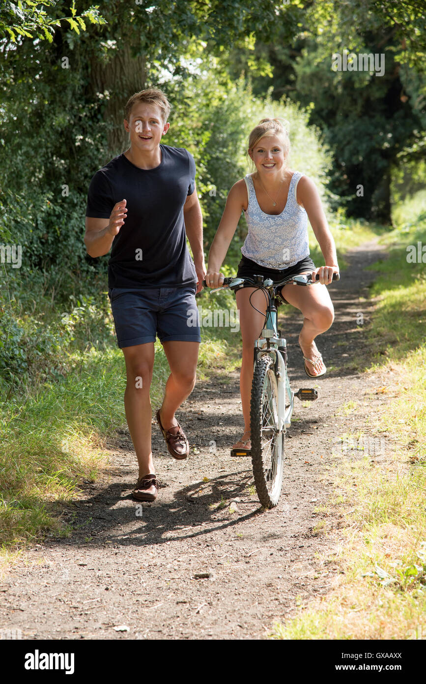 Teenage girl riding a bicycle - Teenager boy running with his female partner riding a bicycle along an English country lane Stock Photo