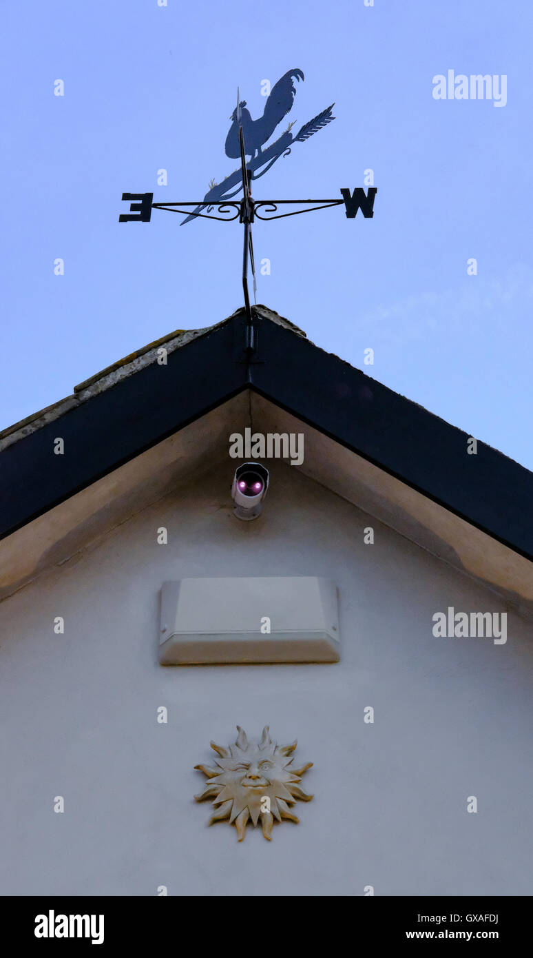 Weather vane, CCTV camera and alarm system seen installed on residential property, with the CCTV showing infra red illumination. Stock Photo