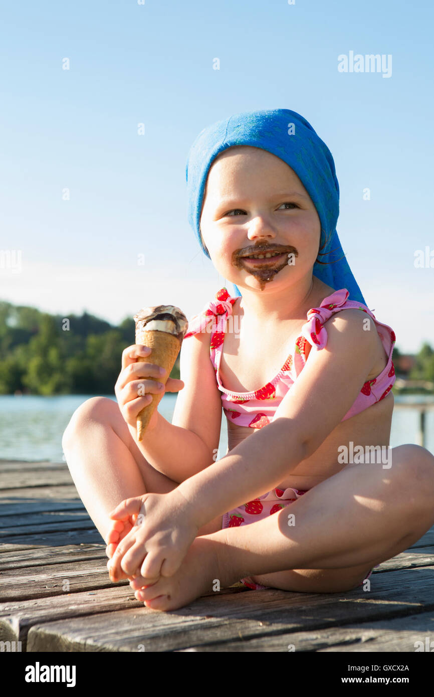 Portrait of female toddler on pier eating chocolate ice cream cone, Lake Seeoner See, Bavaria, Germany Stock Photo