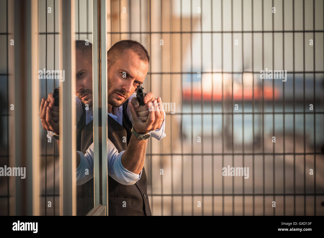 Man in business attire poised behind corner in harbour pointing handgun, Cagliari, Sardinia, Italy Stock Photo