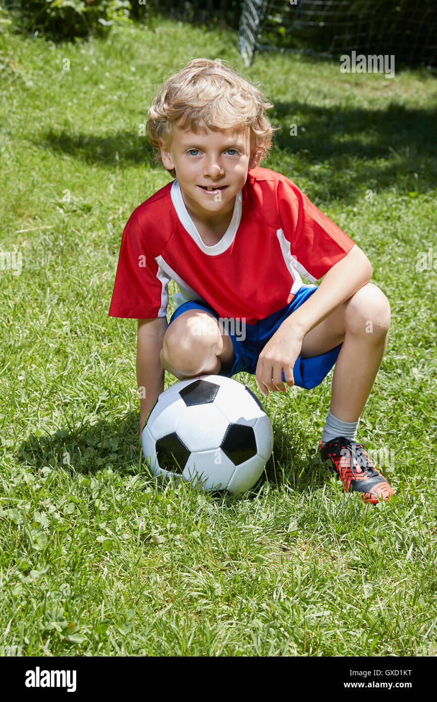 Portrait of boy wearing soccer uniform crouching with soccer ball in garden Stock Photo