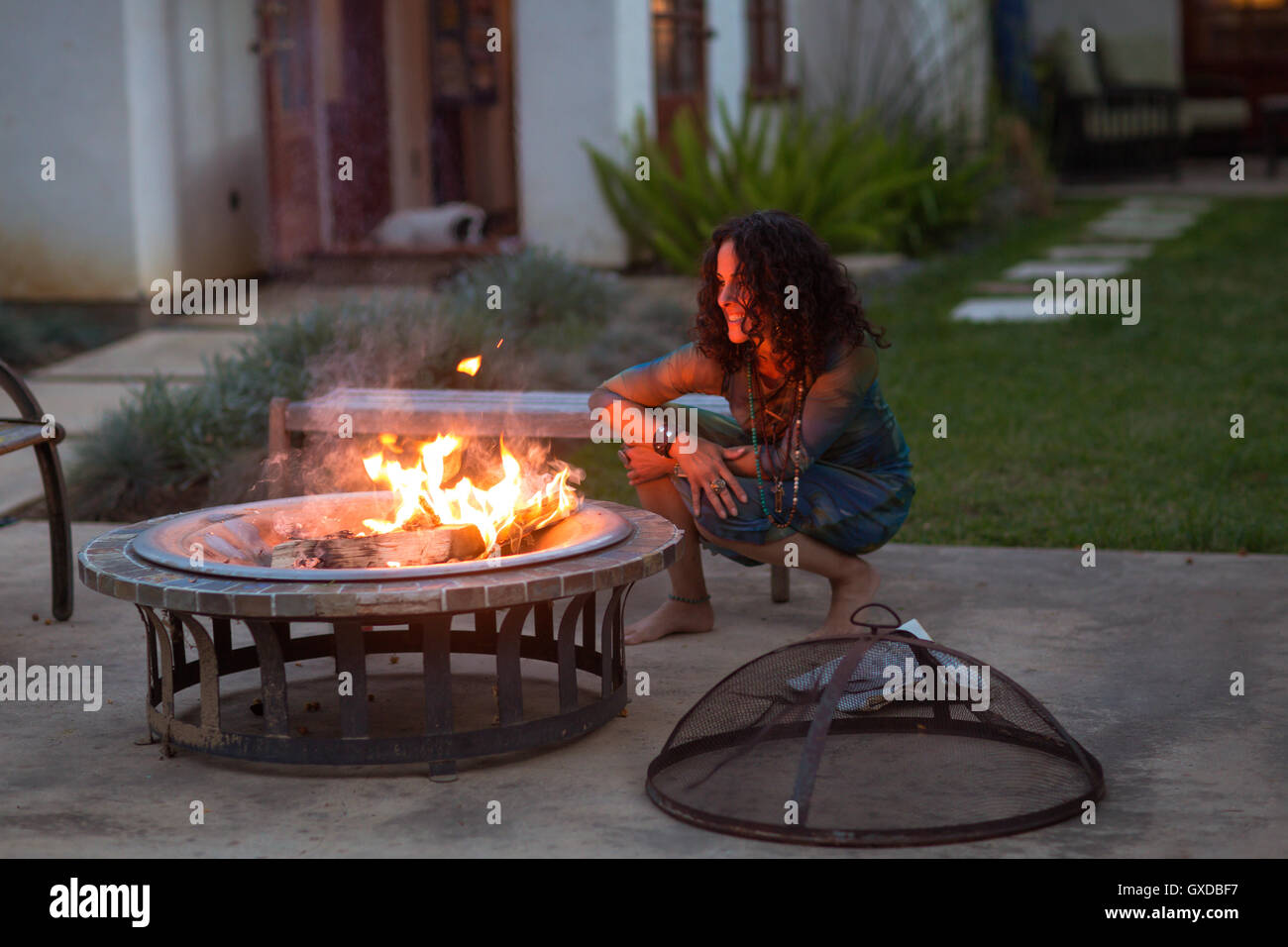 Mature woman crouching in front of patio fire at dusk Stock Photo