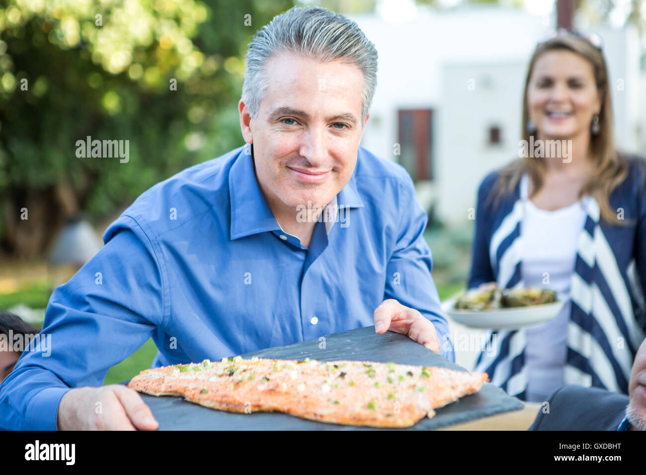 Mature man holding chopping board with fish cuisine at garden party Stock Photo