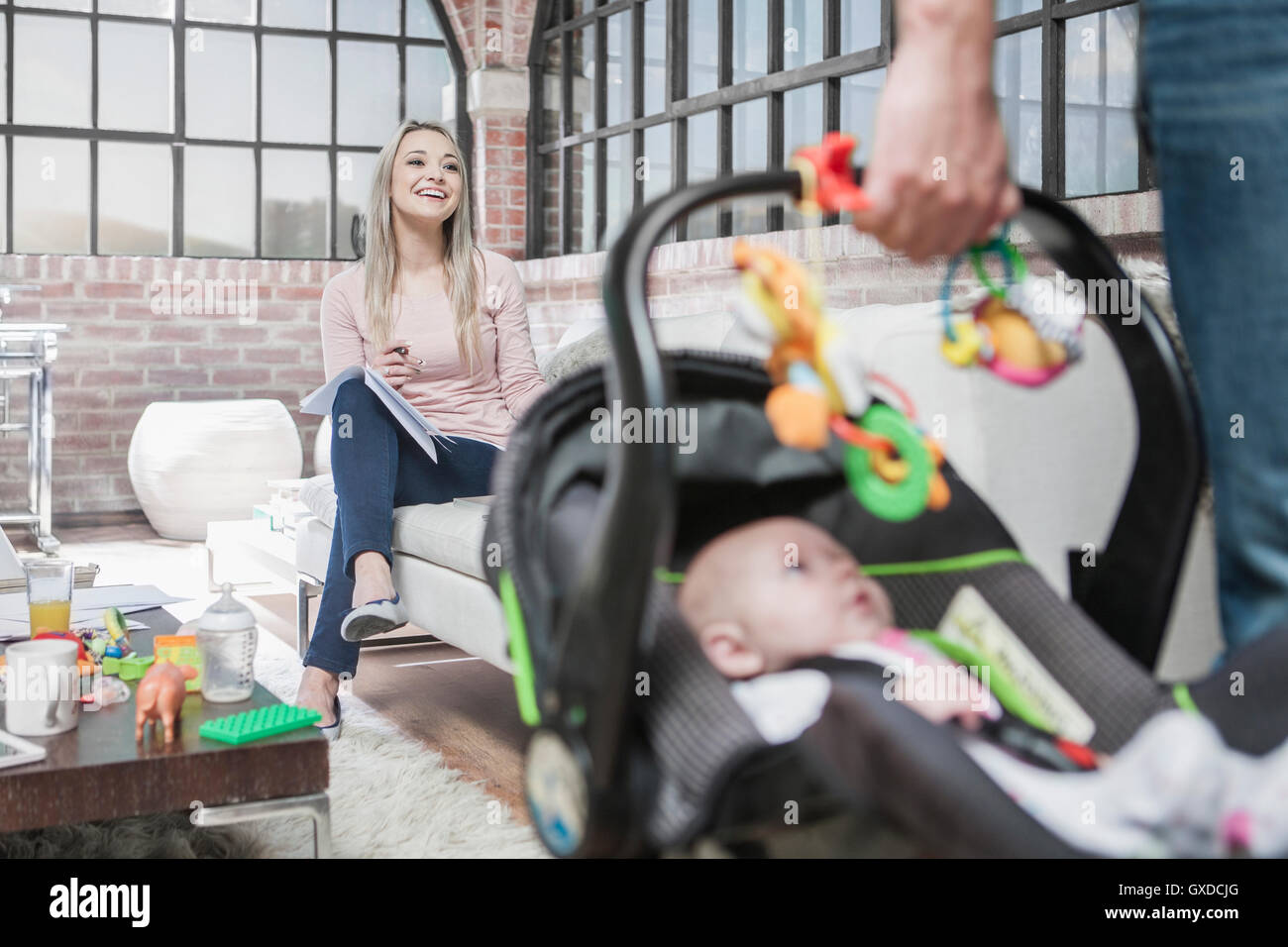 Father leaving house with baby girl in car seat, mother sitting on sofa