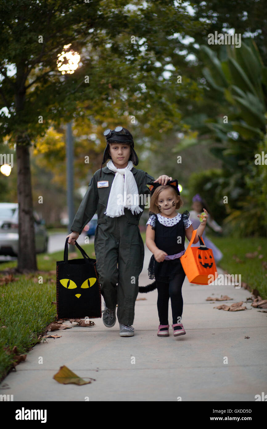 Boy and sister trick or treating in cat and pilot costume walking along sidewalk Stock Photo