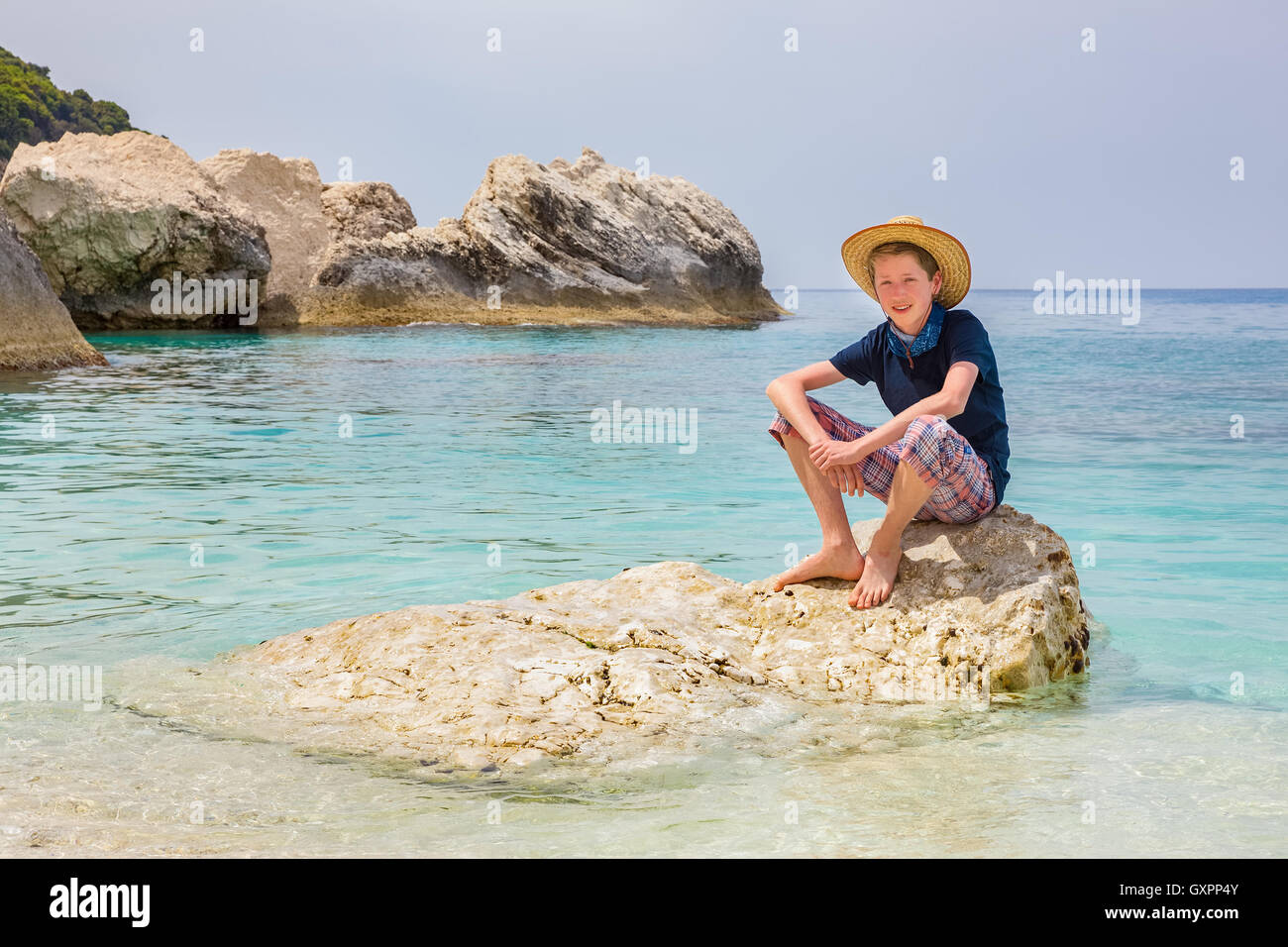 Caucasian teenage boy with hat sitting on rock in sea water Stock Photo