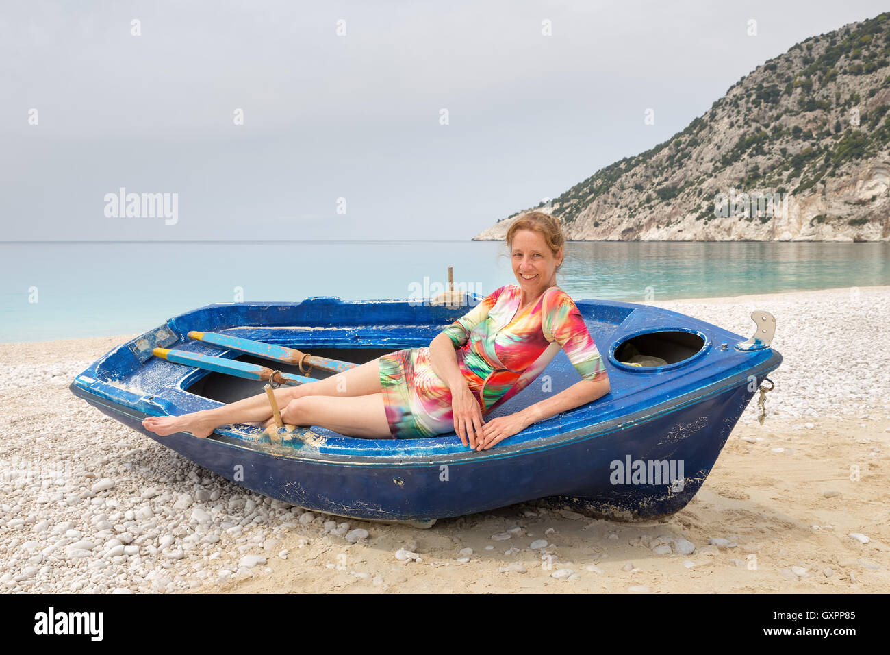 Dutch woman lying in rowing boat on greek beach with mountain and sea Stock Photo