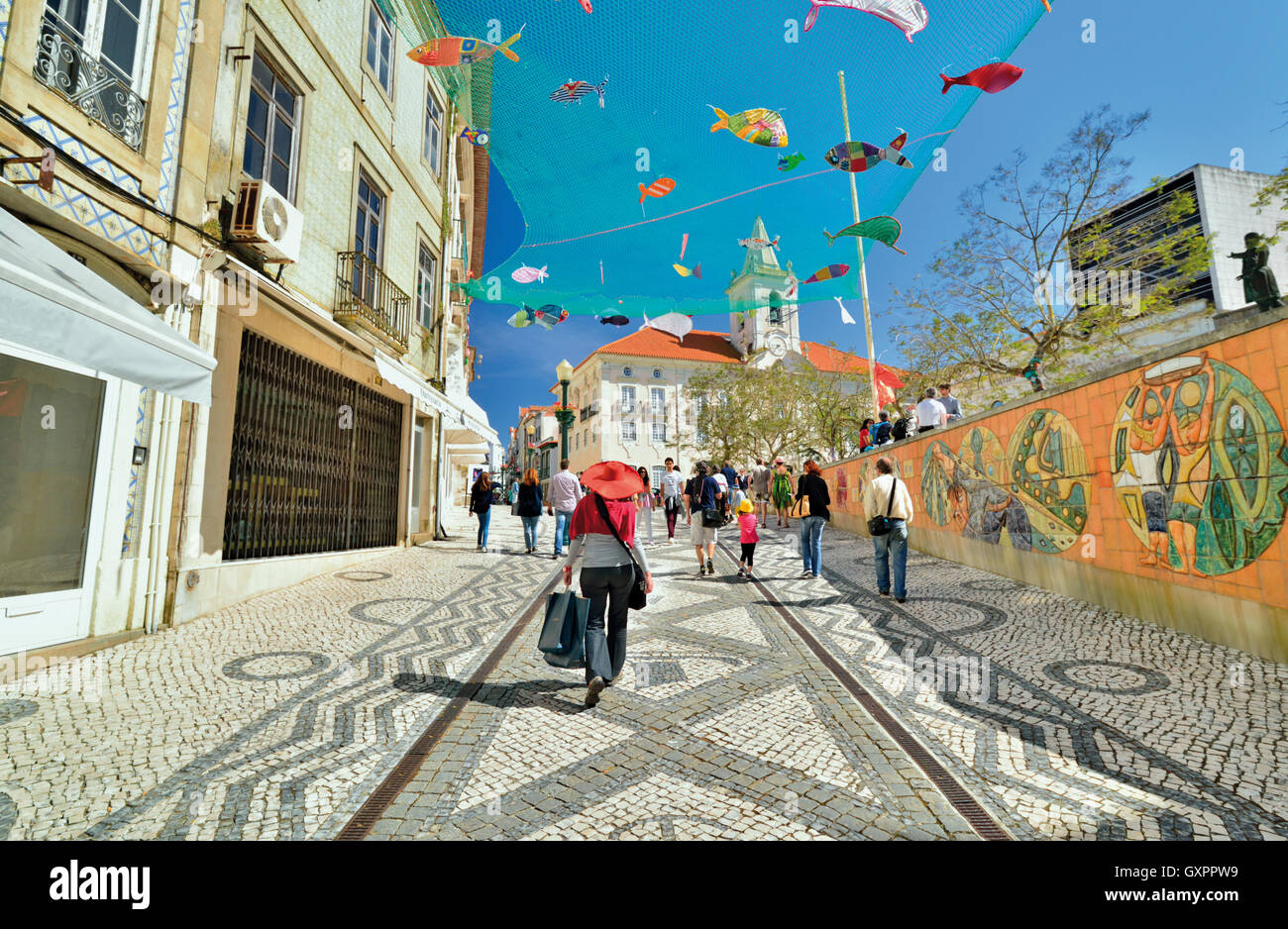 Portugal: Woman with shopping bags and red summer hat walking along decorated cobble stone street with with fish art in the sky Stock Photo