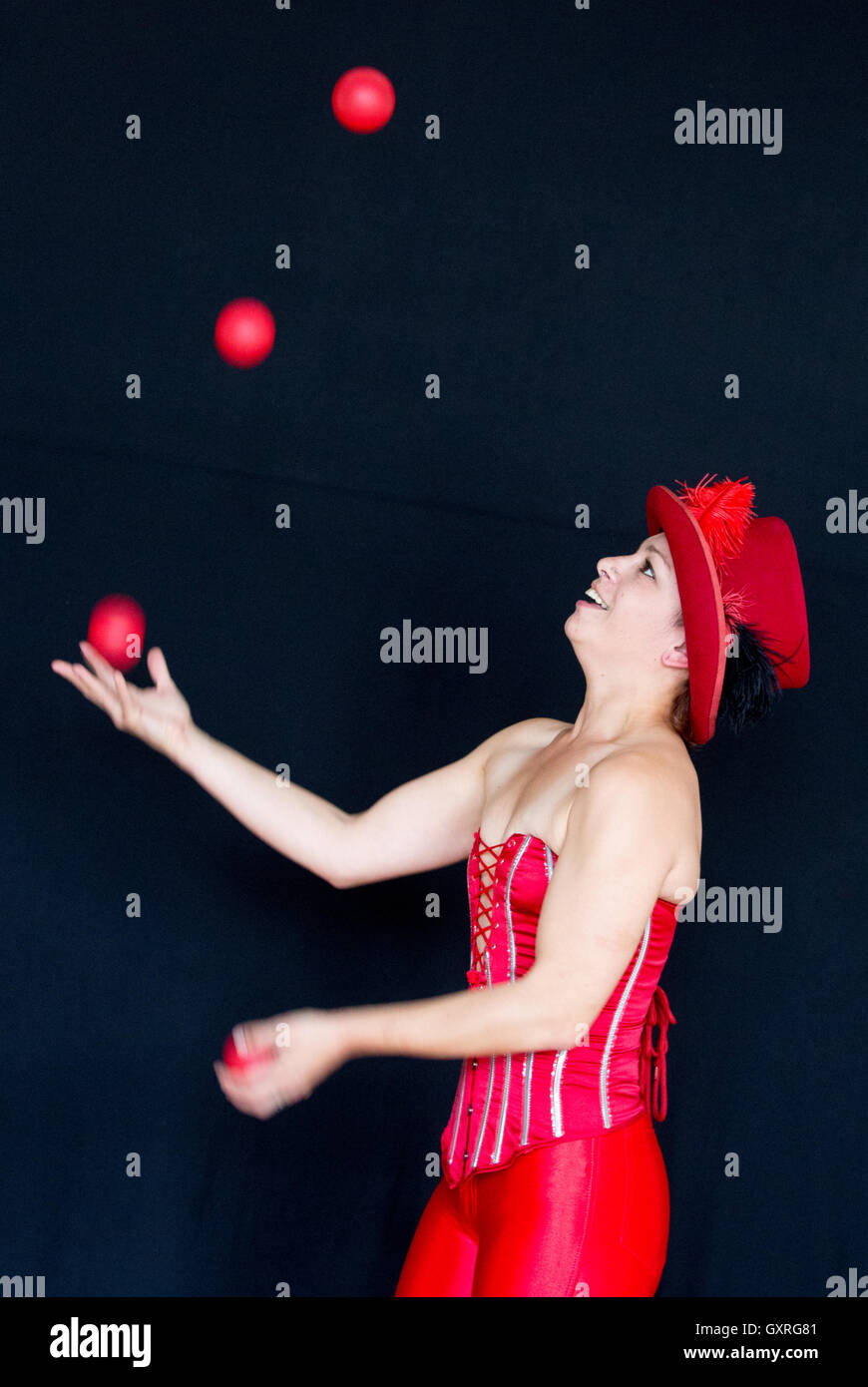 Smiling female juggler wearing a red top hat and red basque with three balls in the air and one in the hand Stock Photo