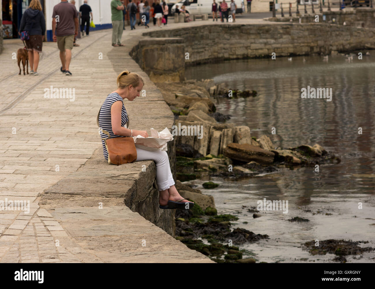 Woman eating fish and chips out of paper while sitting on the edge of a sea wall, Swanage, Dorset, UK. Stock Photo