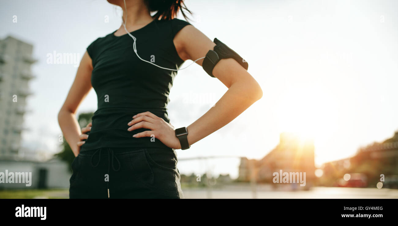 Cropped shot of urban runner outdoors in sunny morning. Fit young woman with smartwatch and mobile phone armband standing with h Stock Photo