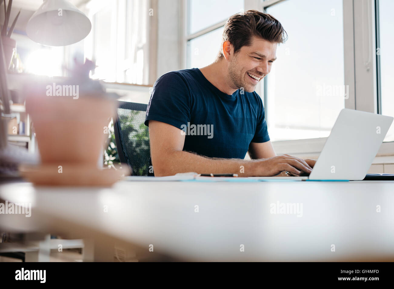 Happy young businessman using laptop at his office desk. Young male executive working on laptop at his desk. Stock Photo