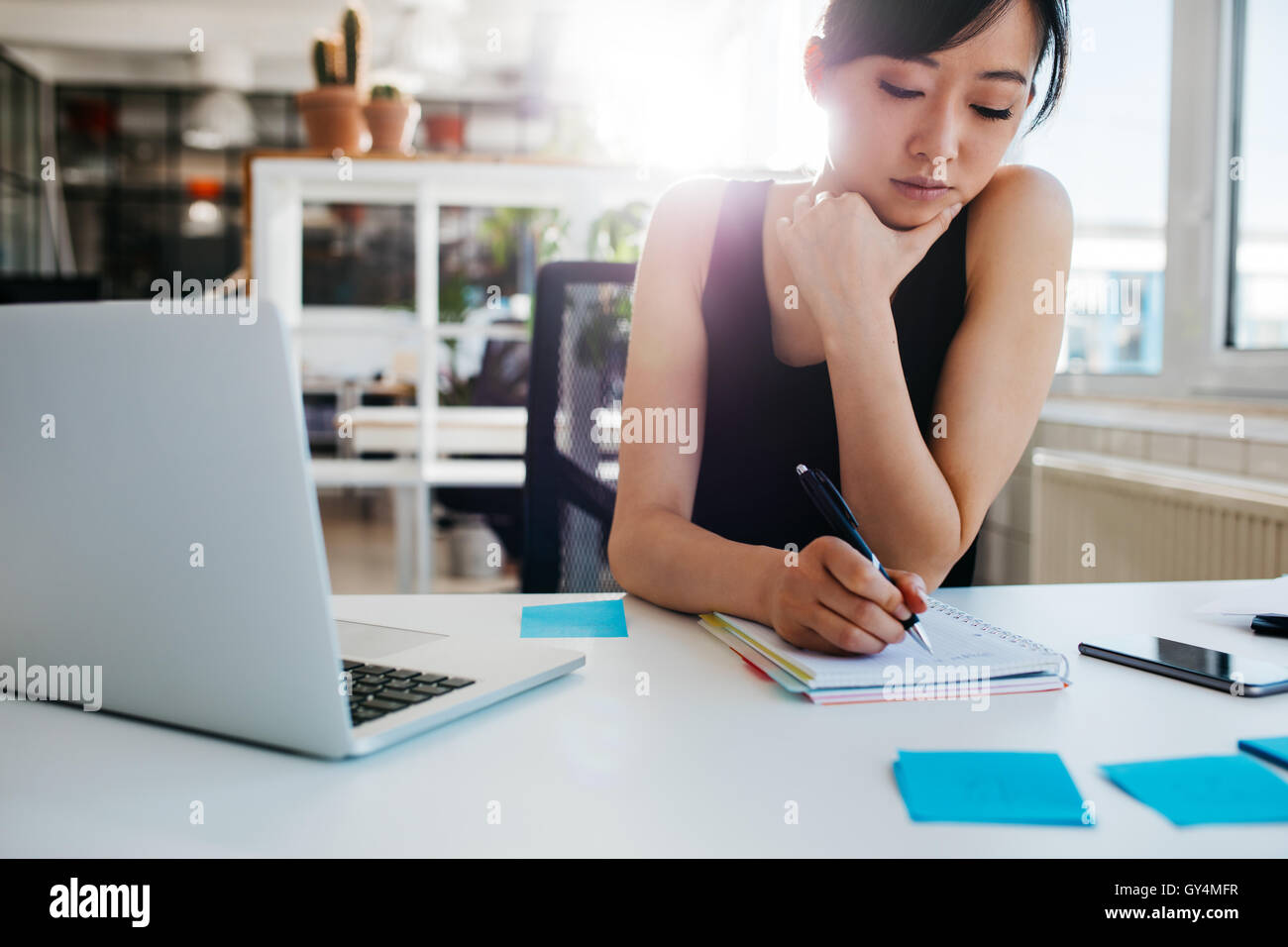 Shot of asian businesswoman writing notes on notepad. Female executive sitting at her desk with laptop taking notes. Stock Photo