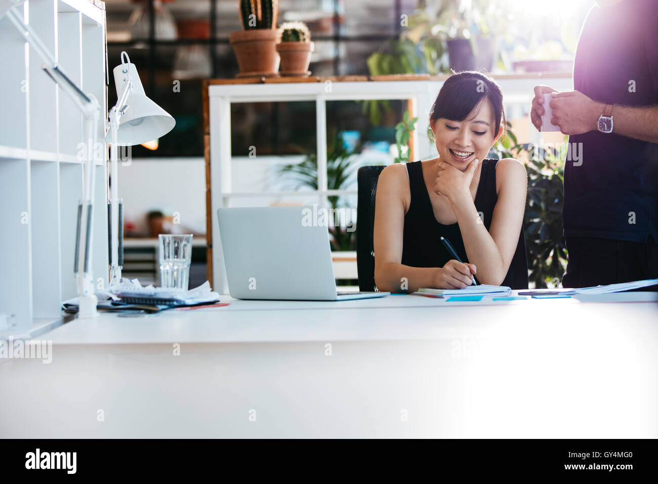 Shot of smiling asian businesswoman writing notes on notepad with colleague standing by. Female executive working at her desk wi Stock Photo