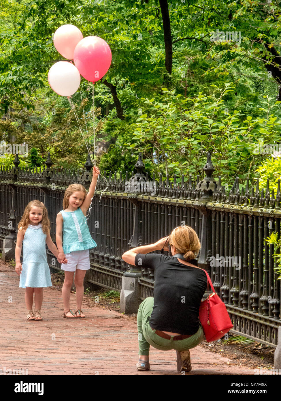 Two sisters hold hands as they pose for a photo by their mother beside a park in Boston, MA. Note balloons and brick sidewalk. Stock Photo