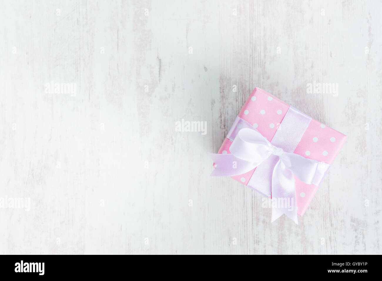 Top view of a gift box wrapped in pink dotted paper and tied satin bow over a white wood background. Stock Photo
