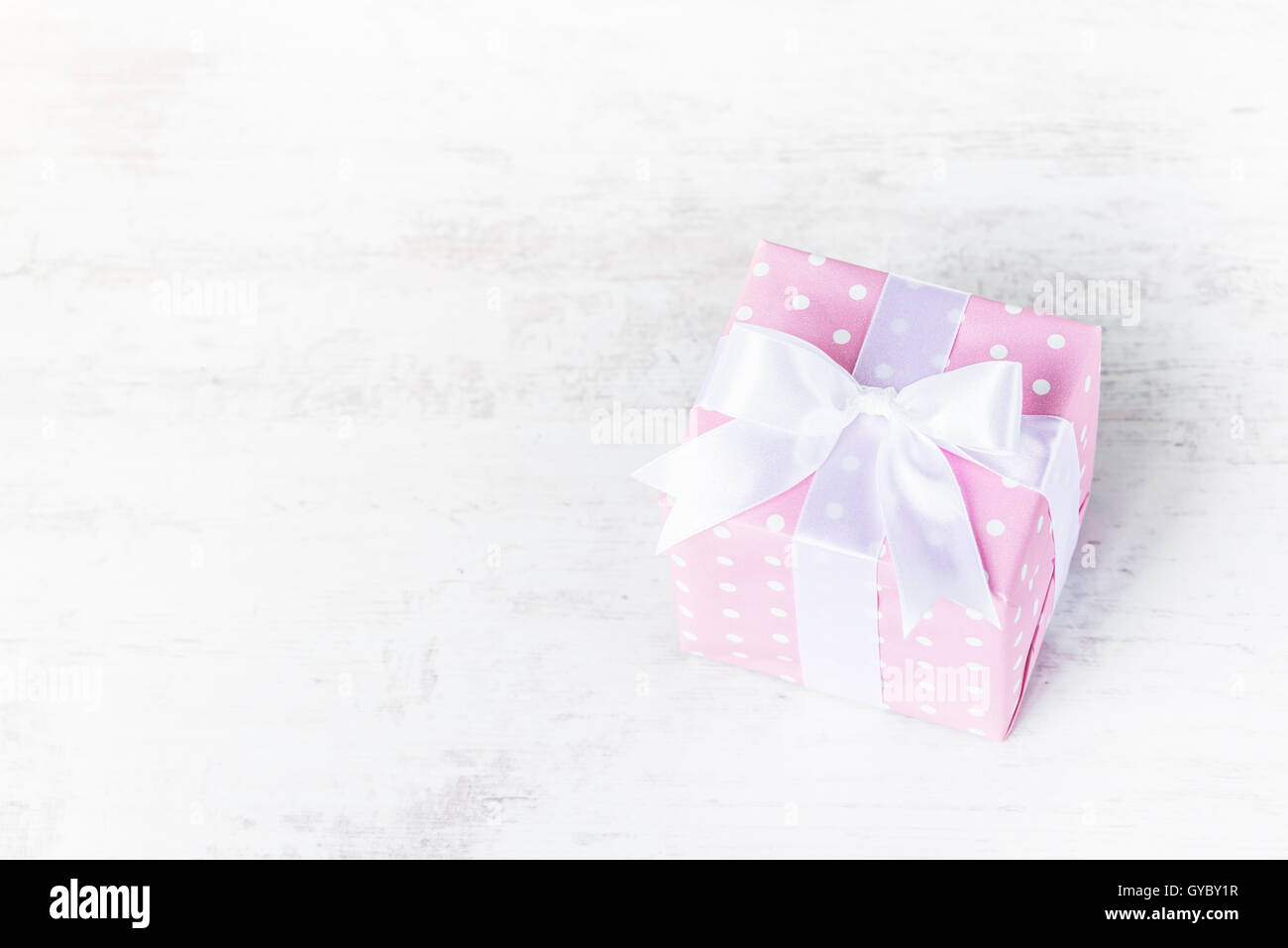 Gift box wrapped in pink dotted paper and tied satin bow over a white wood background. Stock Photo