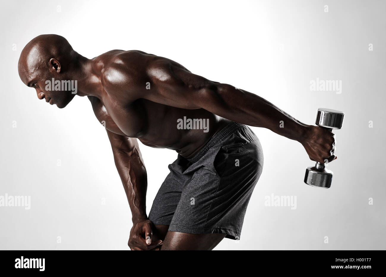 Side view shot of young african man working out with dumbbells against grey background. Muscular man shirtless exercising with h Stock Photo