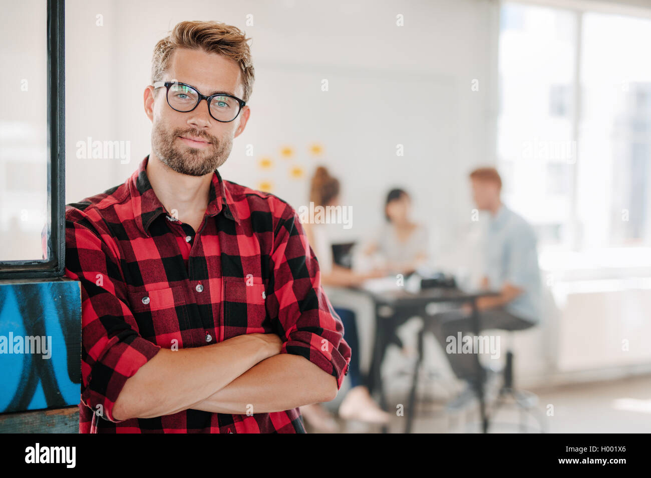 Portrait of a positive looking young business professional standing with his arms crossed with coworkers talking in the backgrou Stock Photo