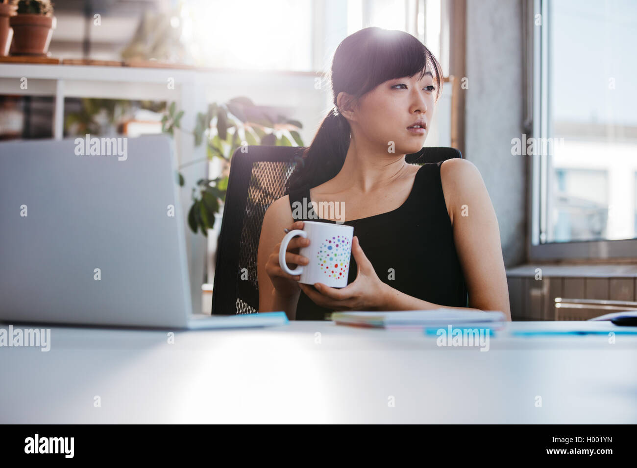 Portrait of relaxed young woman sitting at her desk holding cup of coffee and looking away. Asian business woman taking coffee b Stock Photo