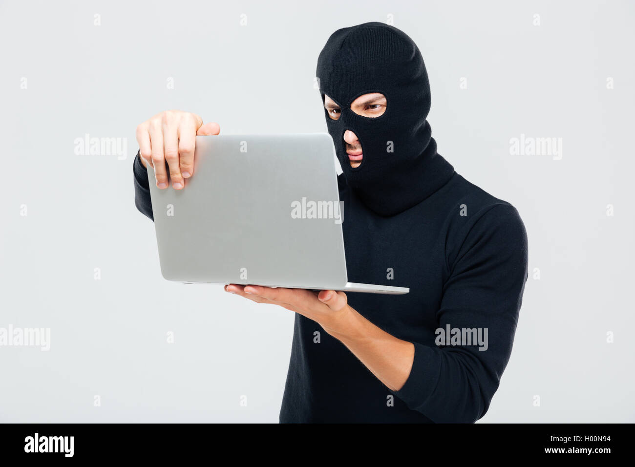 Criminal young man in balaclava standing and using laptop Stock Photo