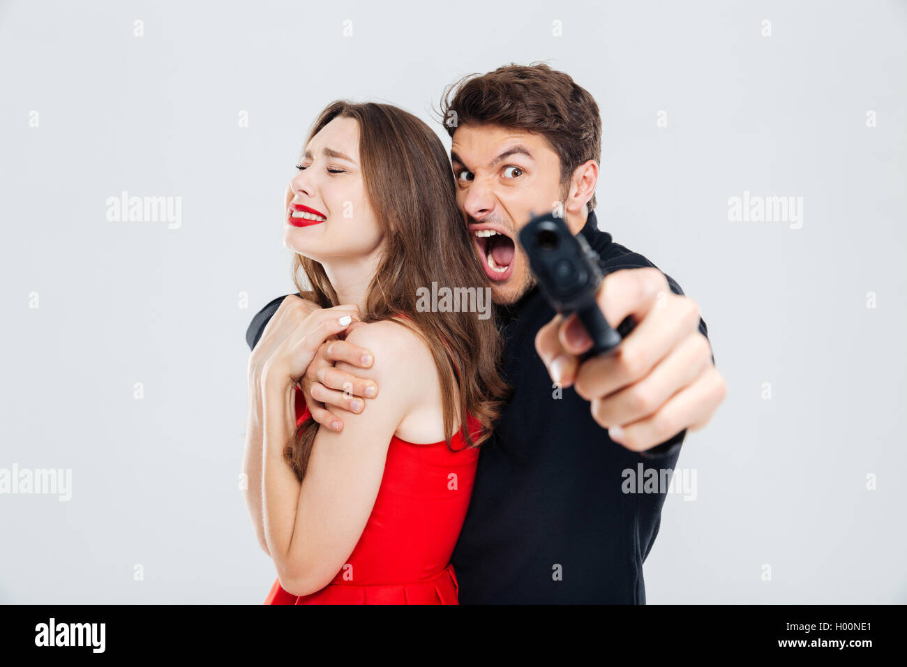 Furious criminal young man holding scared young woman and pointing with gun on you Stock Photo