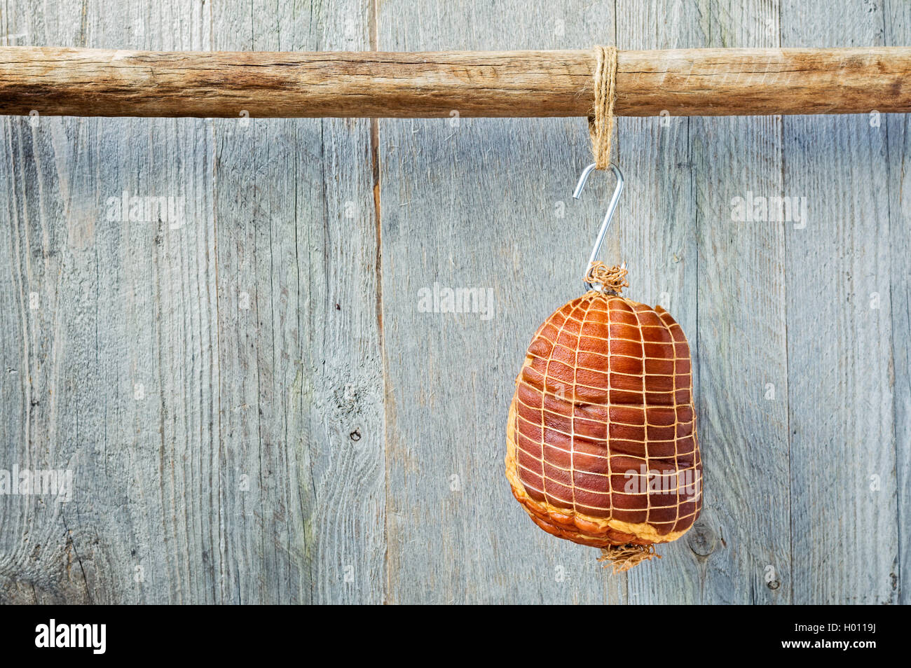Smoked boneless pork ham hock wrapped in netting hanging on a hook from a wooden pole with a weathered wood background Stock Photo
