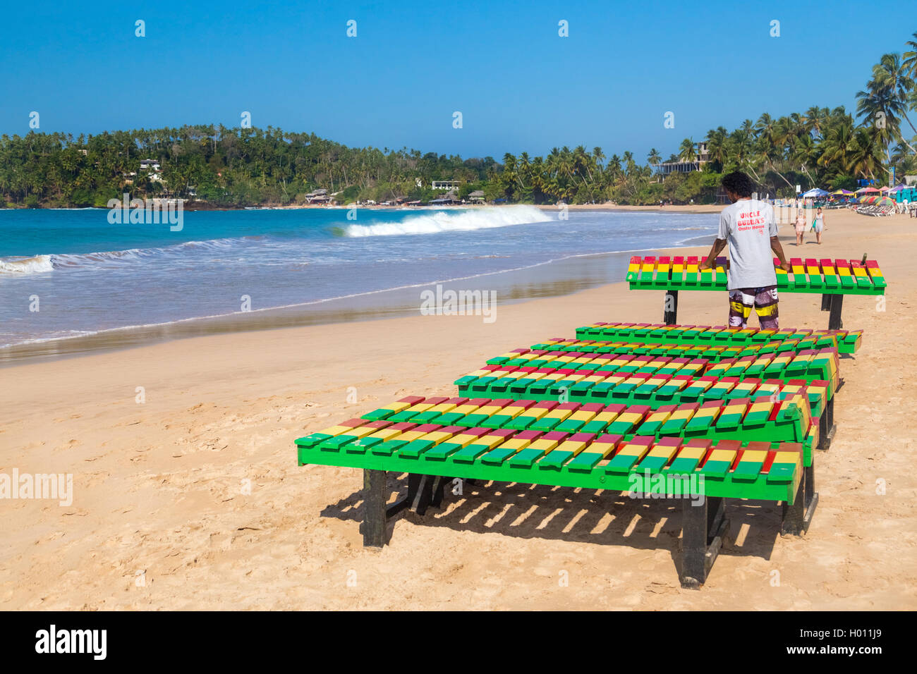 UNAWATUNA, SRI LANKA - MARCH 6, 2014: Local worker putting colourful wooden deck chairs on beach.  Unawatuna is a major tourist  Stock Photo