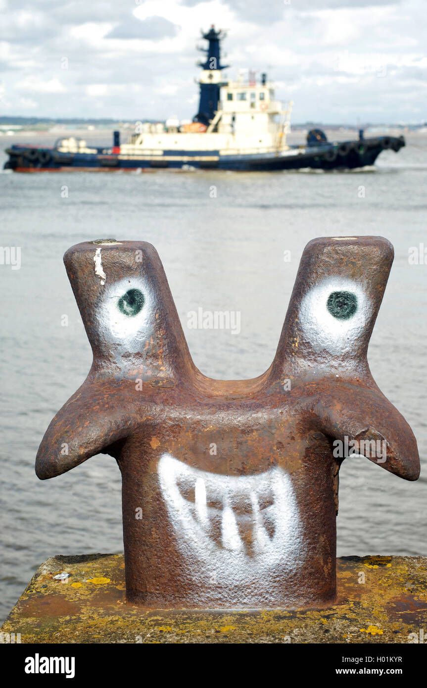 Funny face spray painted on marine mooring bollard on the River Mersey at Liverpool with tug boat passing in the background Stock Photo