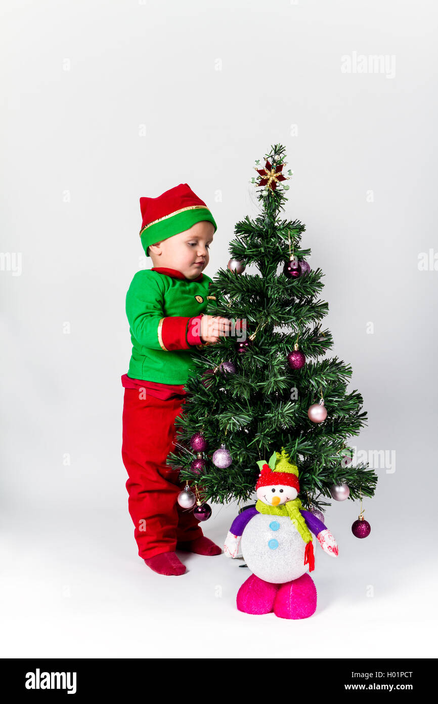 Baby boy dressed as Santa's Helper decorating  Christmas tree, hanging ornaments. White background. Stock Photo
