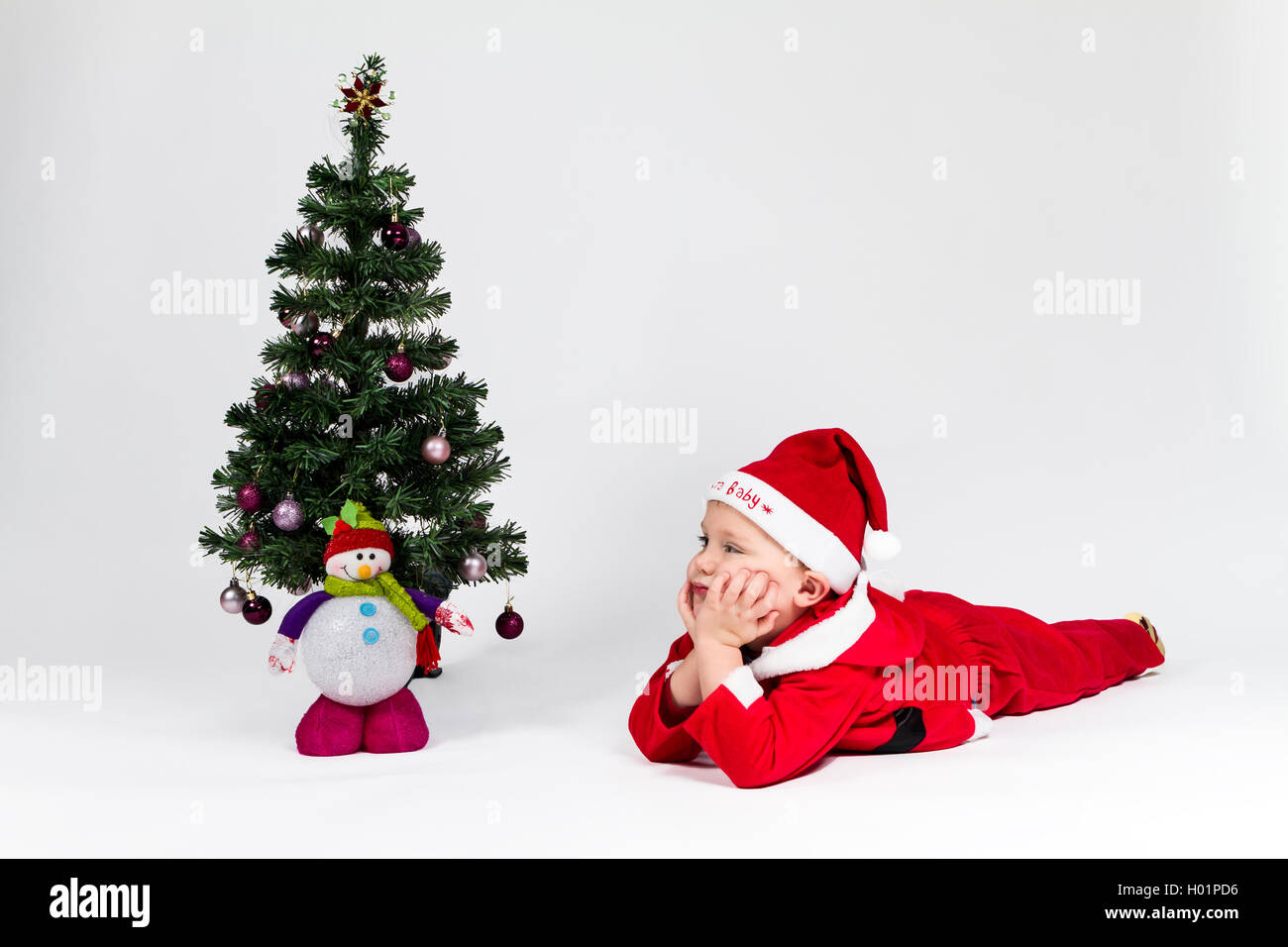 Dreaming baby boy dressed as Santa Claus lying next to Christmas tree. White background. Stock Photo