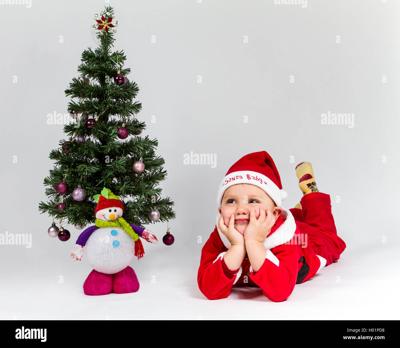 Dreaming baby boy dressed as Santa Claus lying next to Christmas tree. White background. Stock Photo