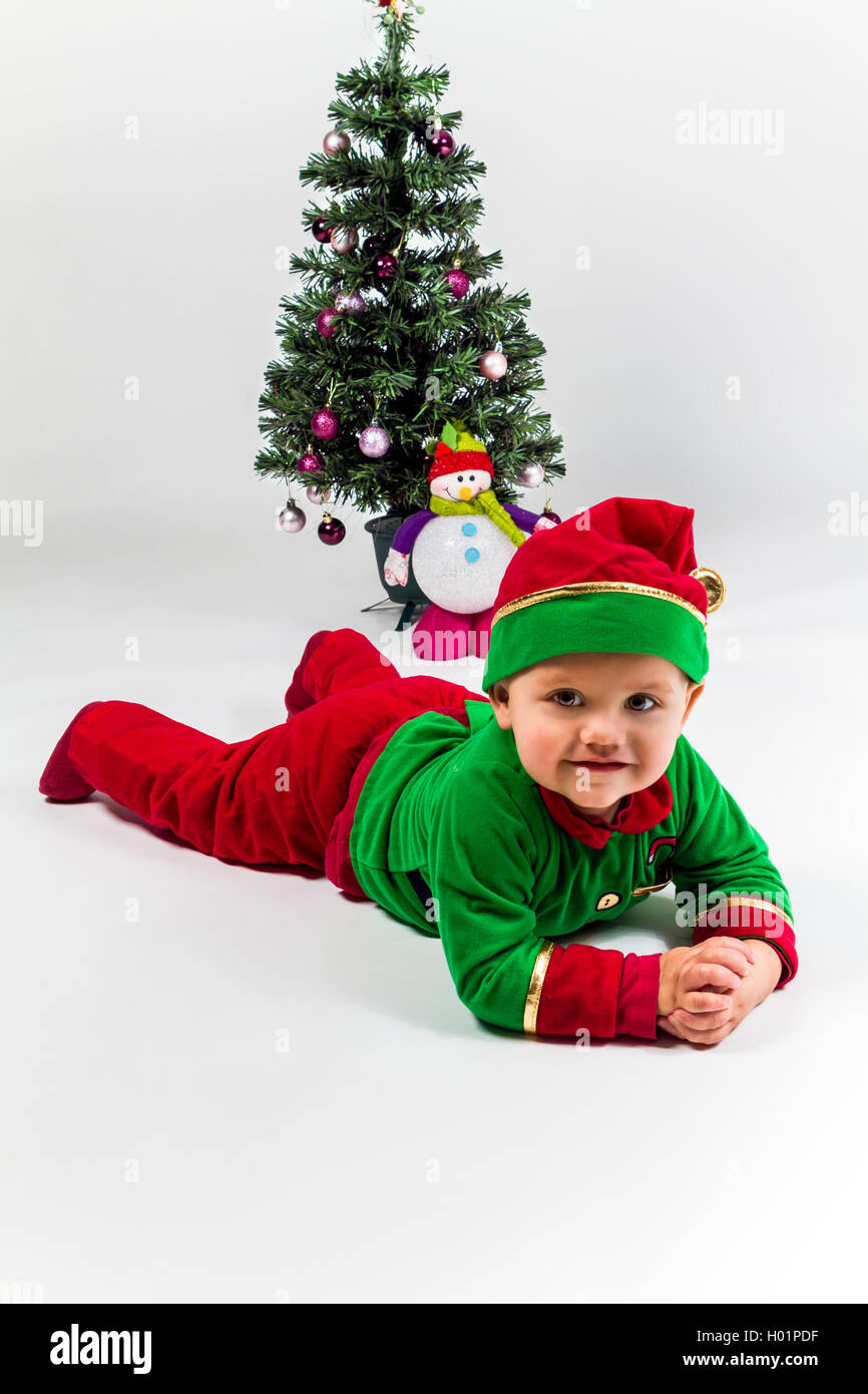 Baby boy dressed as Santa's Helper lying next to Christmas tree. White background. Stock Photo