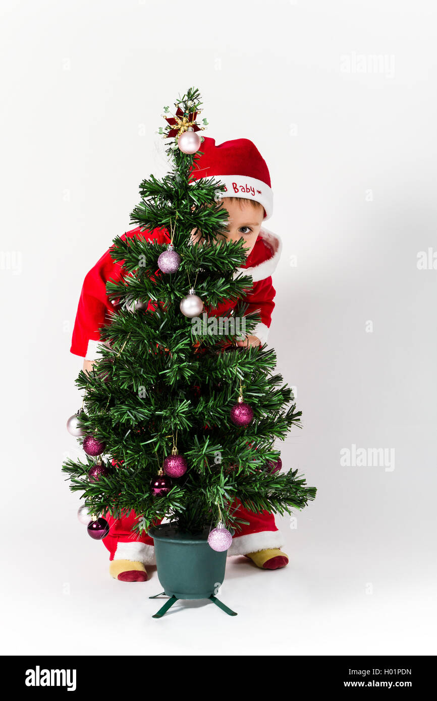 Baby boy dressed as Santa Claus hiding behind Christmas tree. White background. Stock Photo