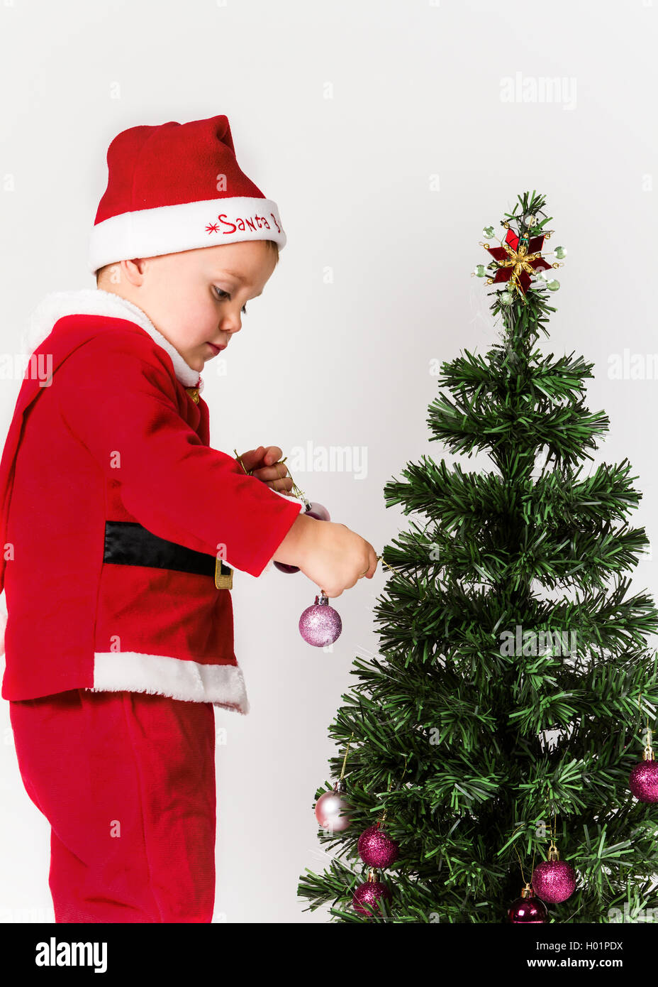 Baby boy dressed as Santa Claus decorating  Christmas tree, hanging ornaments. White background. Stock Photo
