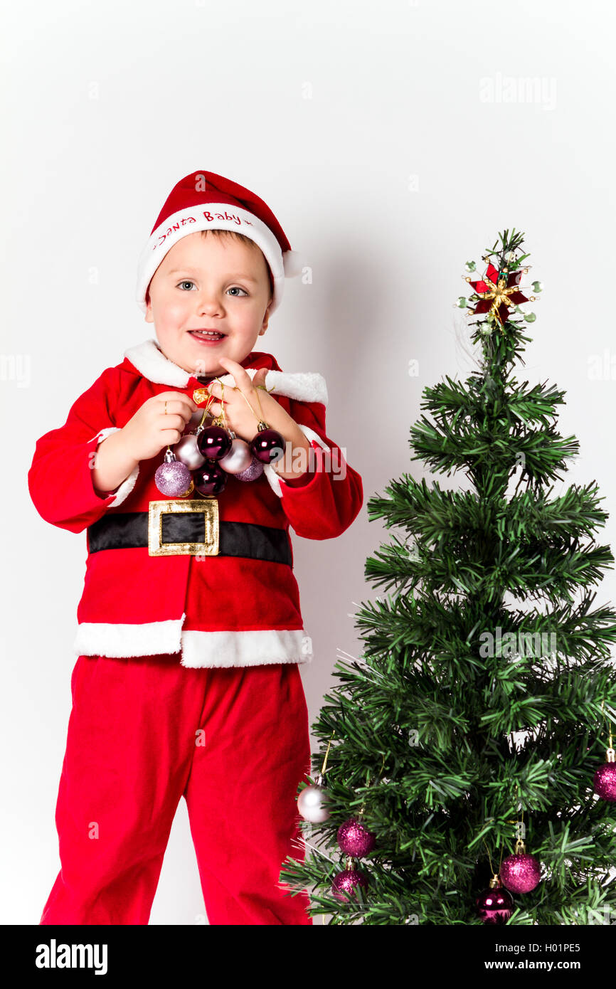 Baby boy dressed as Santa Claus decorating  Christmas tree, holding baubles. White background. Stock Photo