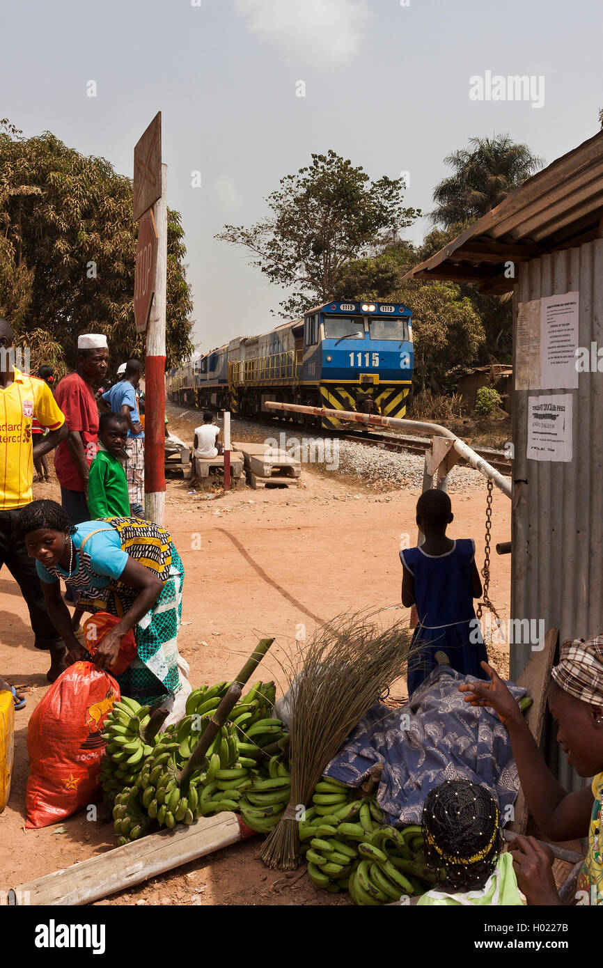 Iron ore project. Village people on market day waiting at train crossing barrier while loaded ore train from mine passes on its way to port Stock Photo