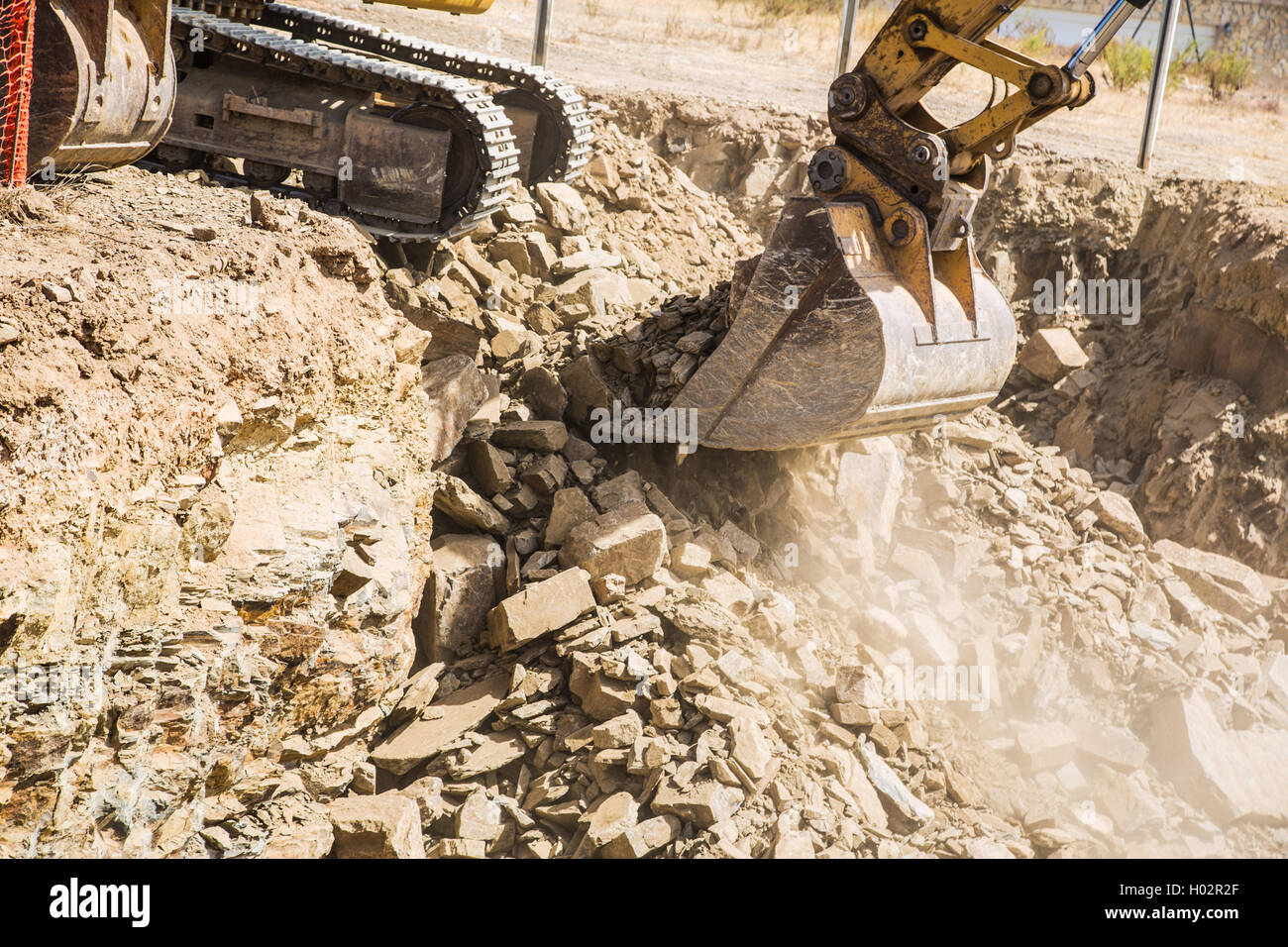 Scoop of an excavator digger removing rocks during a excavation. the tracks of the machine can be seen and dust from the dig. Stock Photo