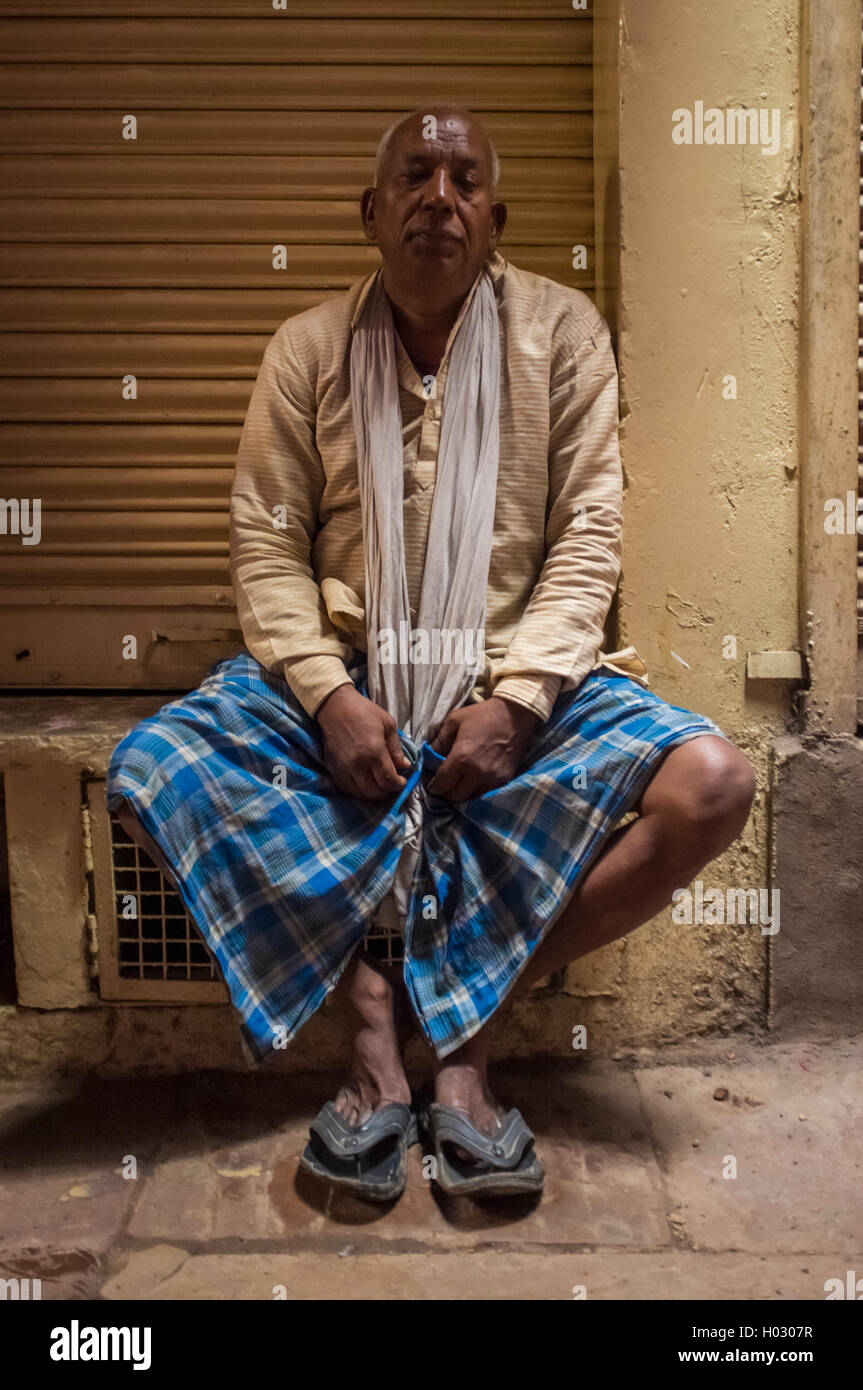 VARANASI, INDIA - 20 FEBRUARY 2015: Indian man sitting next to closed store wearing shirt, scarf, lungi and slippers. Stock Photo