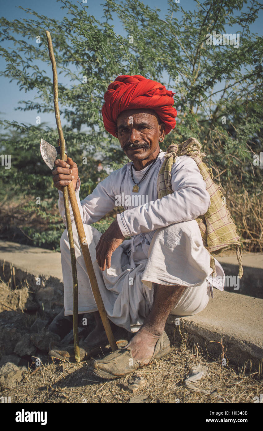 GODWAR REGION, INDIA - 14 FEBRUARY 2015: Elderly Rabari tribesman with red turban sits and holds ax and stick. Post-processed wi Stock Photo