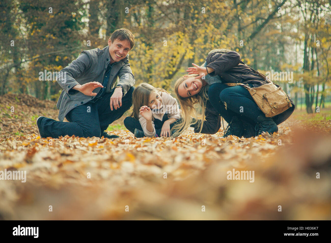 Cheerful family of three kneel on park ground covered with leaves on an autumn day. Stock Photo