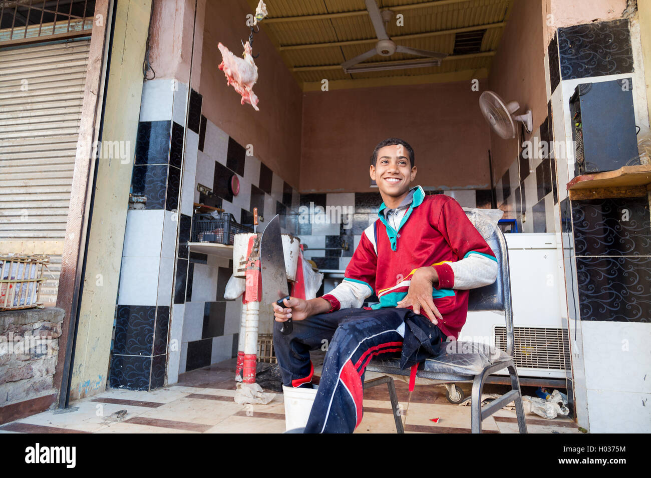 DARAW, EGYPT - FEBRUARY 6, 2016: Young local butcher sitting on the chair holding knife in front of butcher shop. Stock Photo