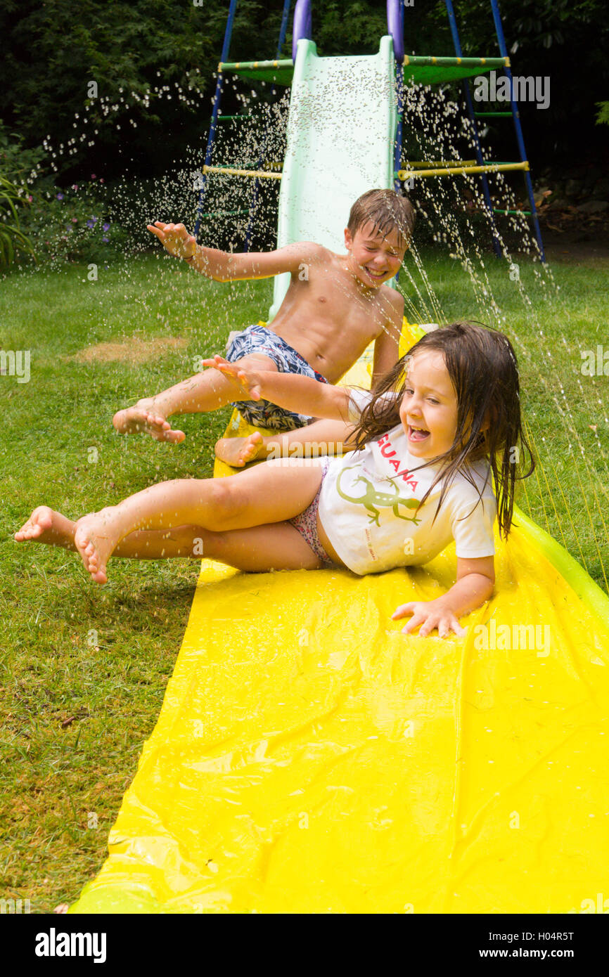 Brother and sister, ten and three years old, playing together on water slide in back garden. UK