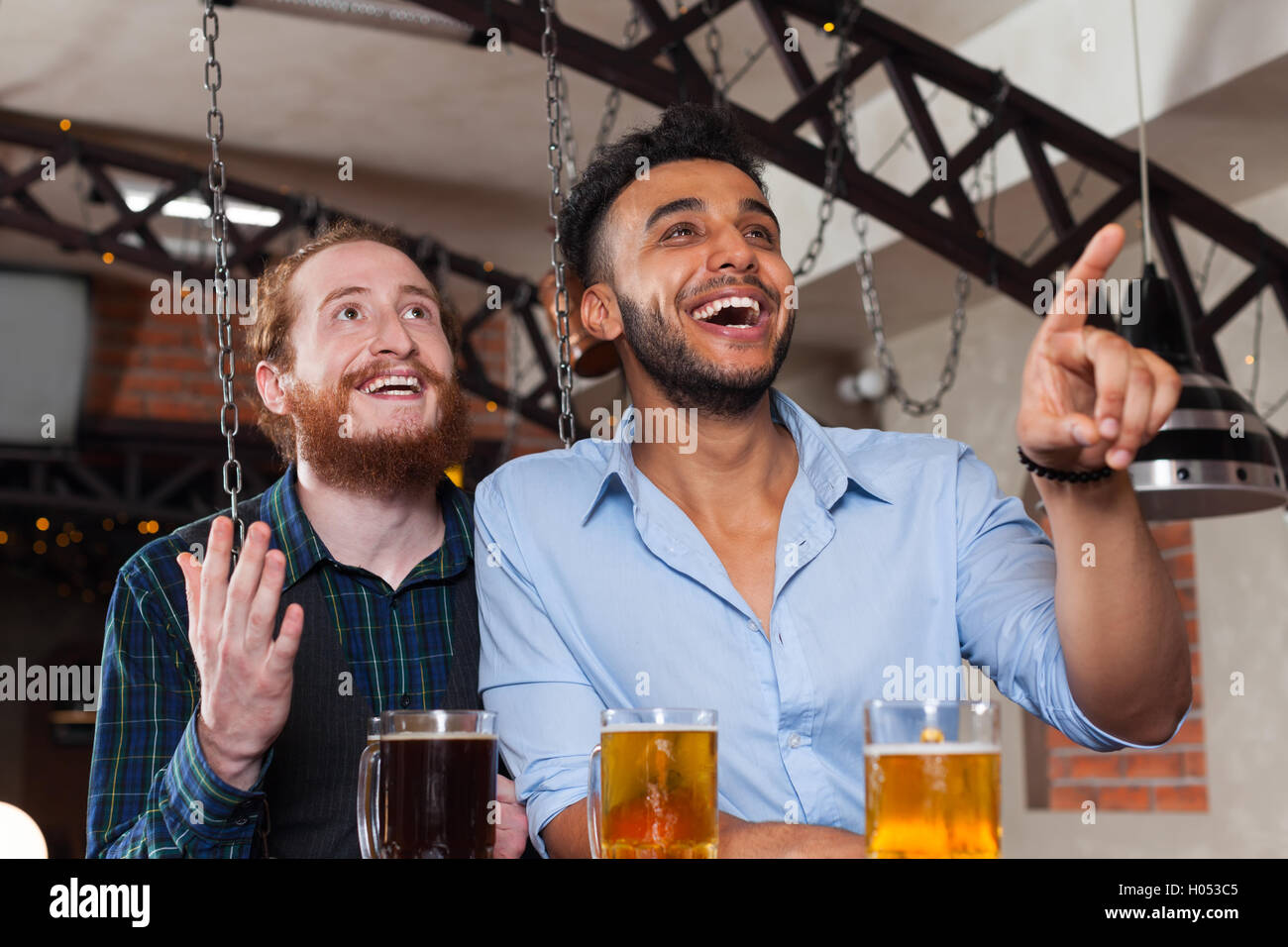 Two Man In Bar Watching Football, Drinking Beer Standing At Counter Point Finger, Mix Race Cheerful Friends Stock Photo